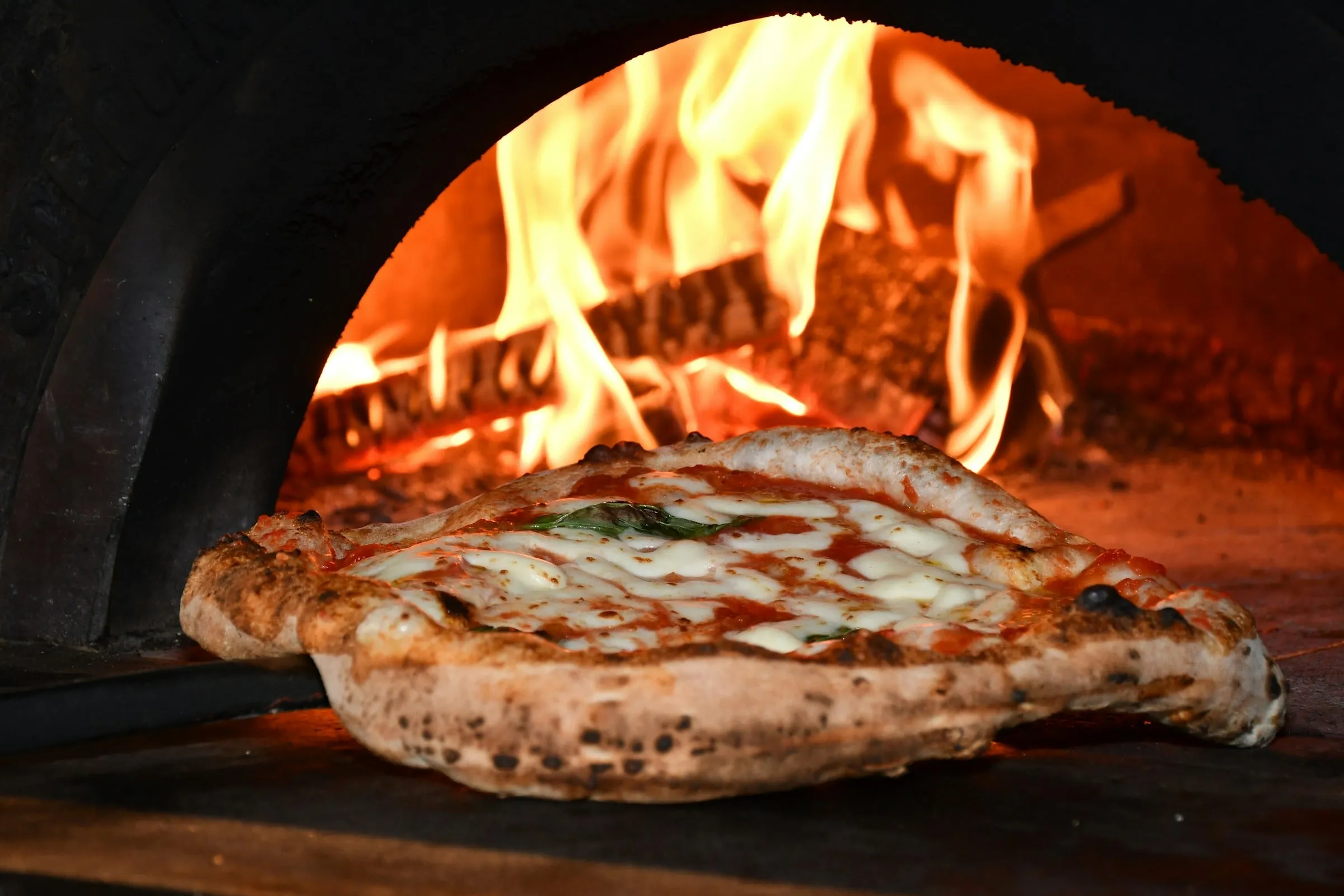 A pizza on a wooden peel inside a brick oven with flames behind it
