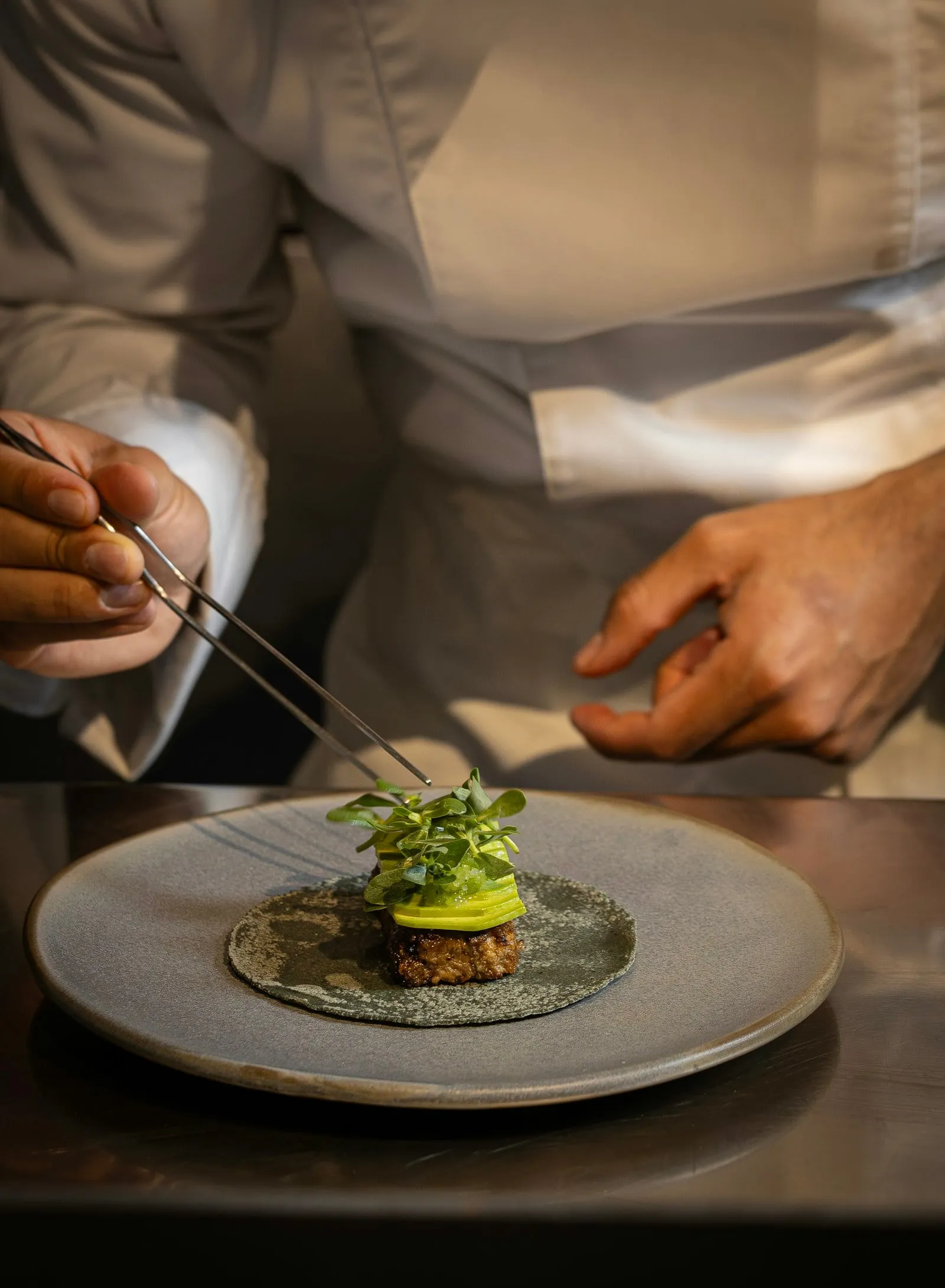 A chef in whites placing herbs on a plate with tweezers