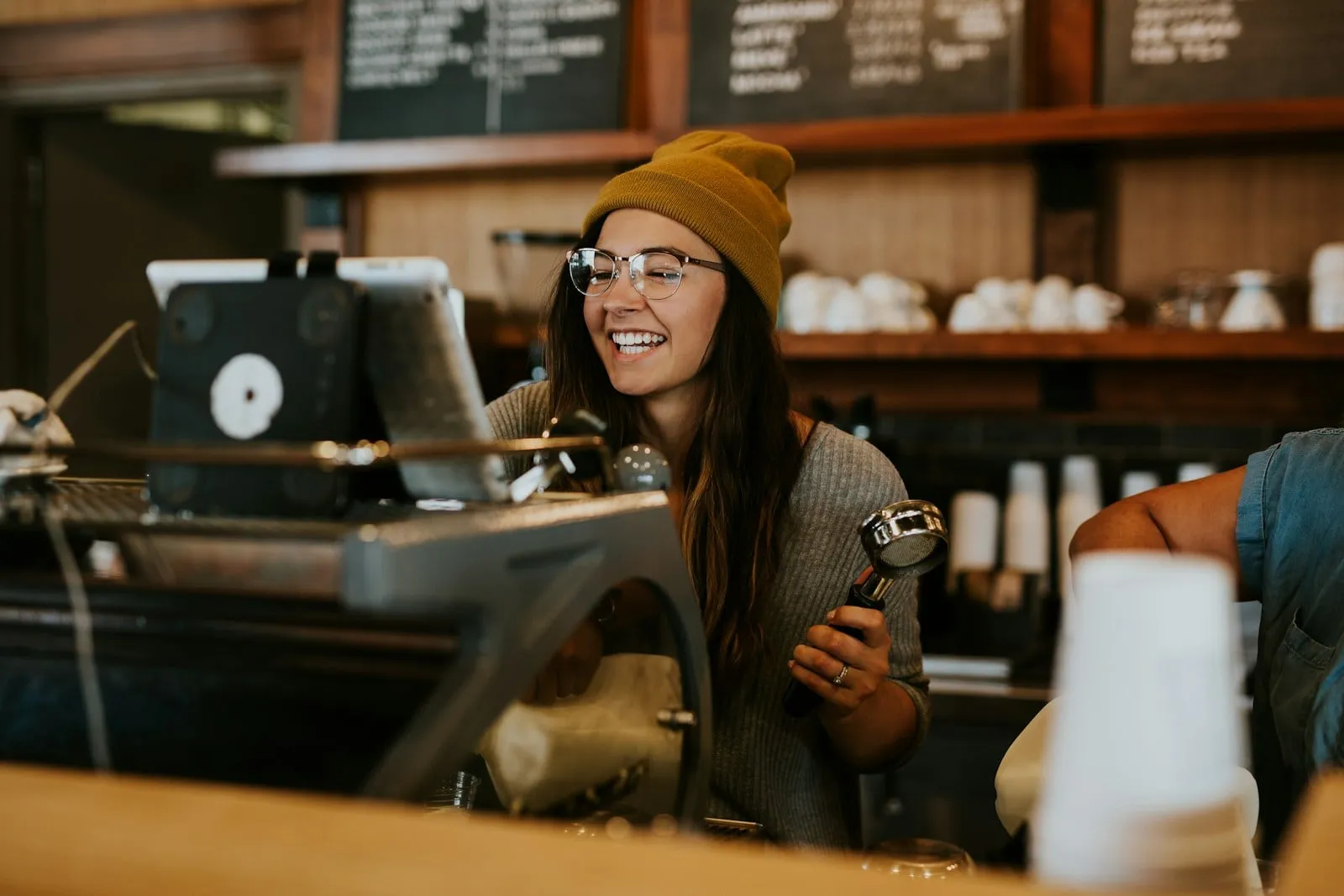A smiling barista in a yellow beanie behind the espresso machine
