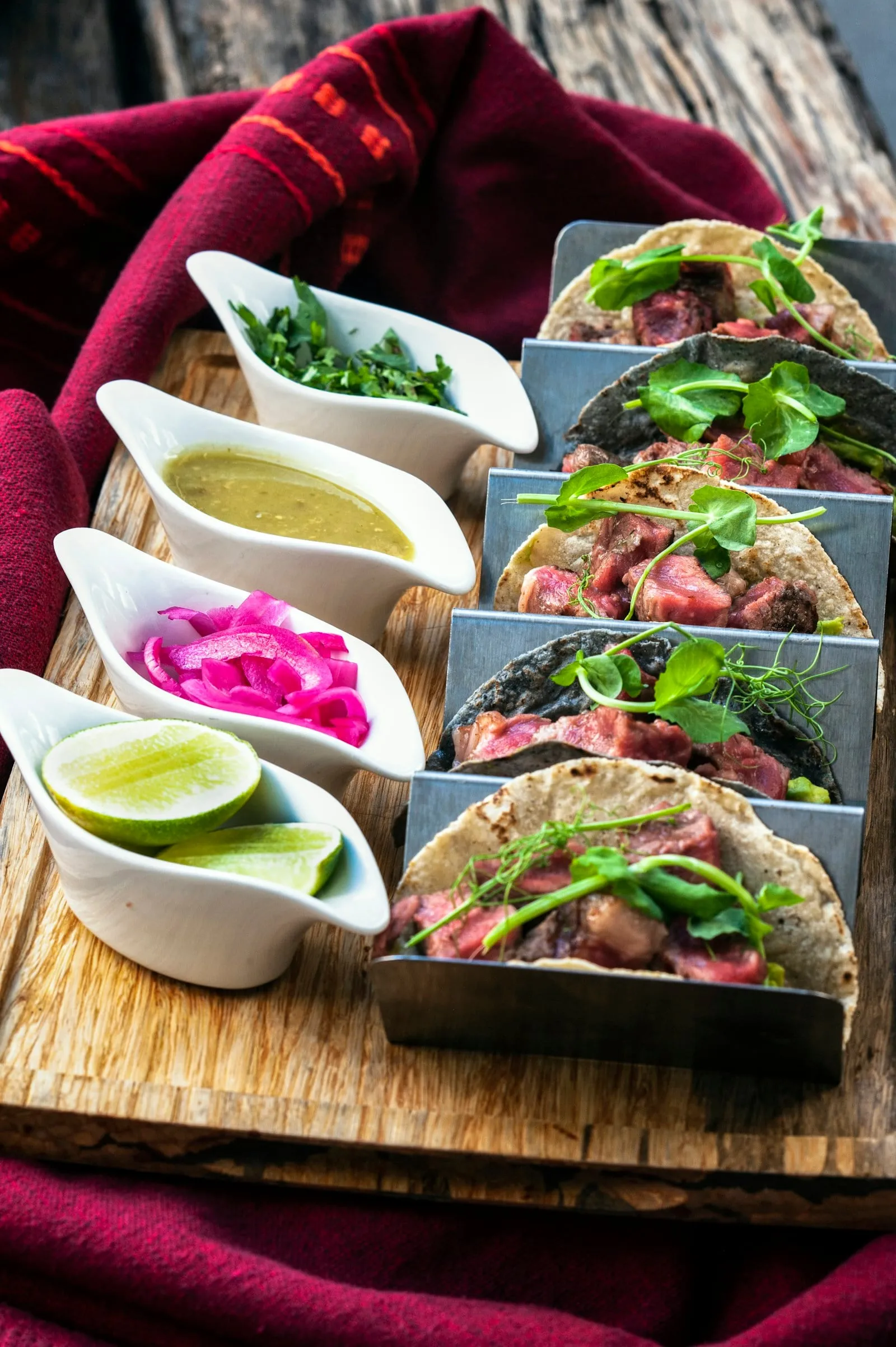 A row of tacos with salsas and limes on a wooden board