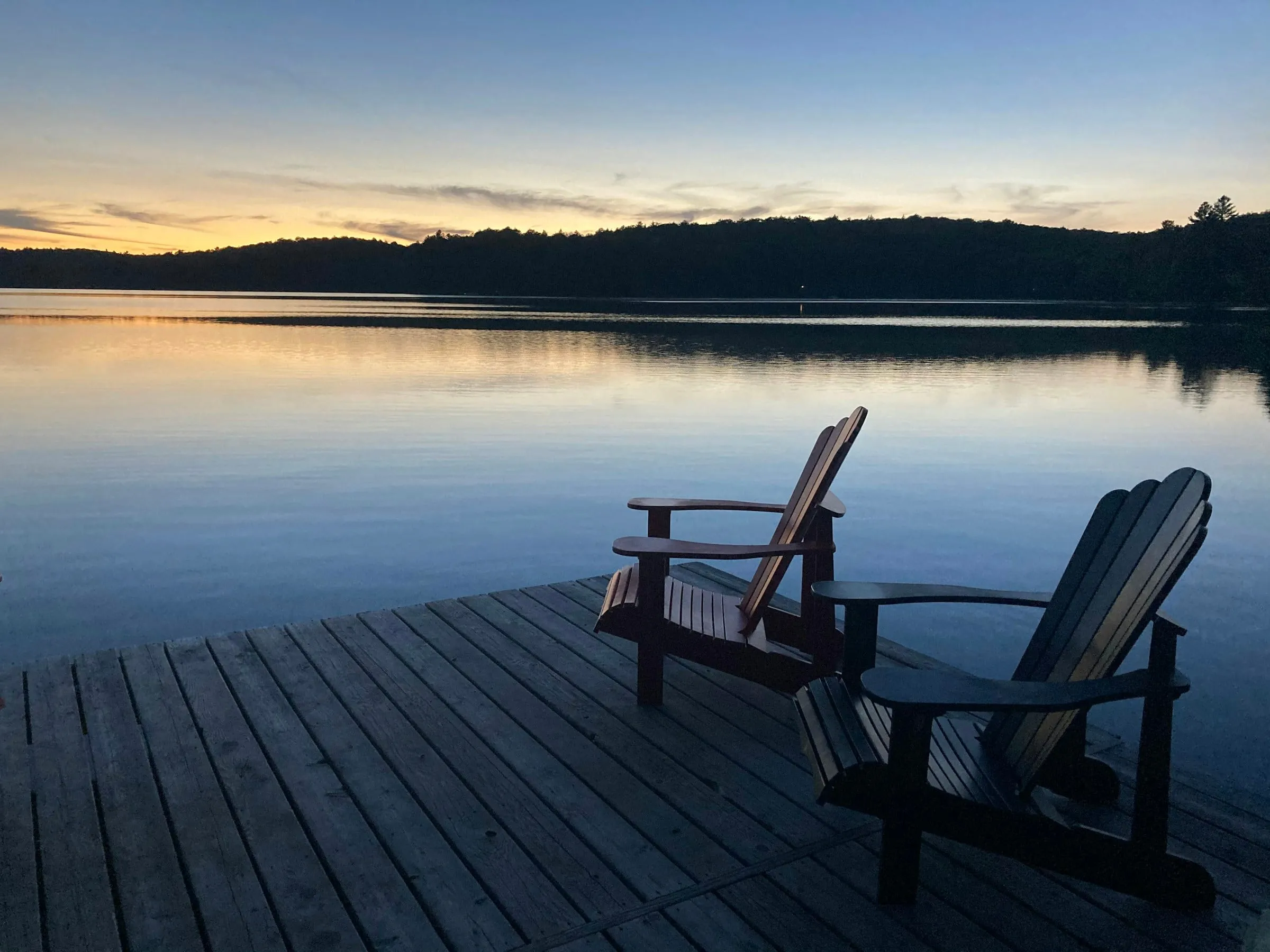 Two wooden deck chairs on a dock looking out over a calm lake at dusk