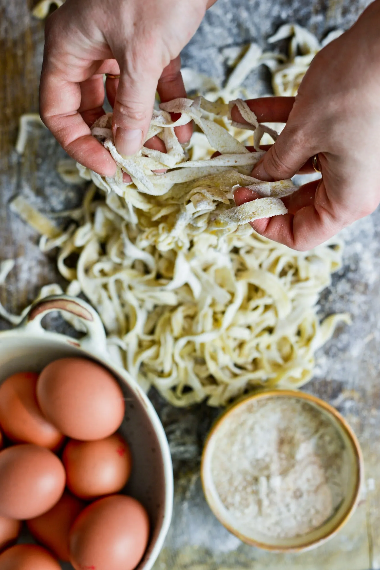 Hands working fresh egg pasta beside a bowl of eggs and flour